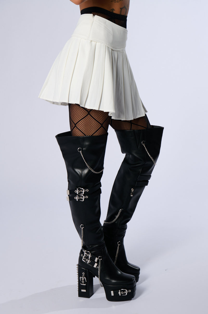 A person is wearing the SHAKE IT UP PLEATED MINI SKIRT in white, paired with black fishnet stockings and black thigh-high platform boots adorned with silver chains and buckles. They are posing against a plain white background.