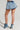 A person stands against a neutral background in a stylish ensemble featuring the BELLA BABY DENIM MINI SKIRT with built-in shorts, perfectly complemented by matching high-heeled denim boots, showcasing their chic fashion style.