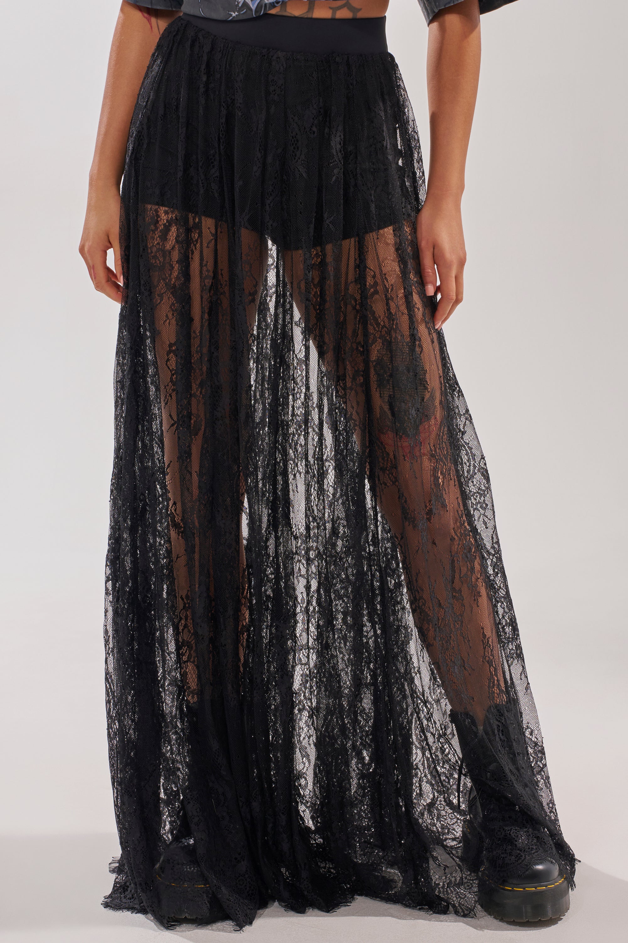 A person wears the LOVELY DAYS LACE MAXI SKIRT over black shorts with lace-up boots—an effortlessly chic resort look. The mid-torso-down crop highlights the skirt’s sheer black lace and stylish details. 0
