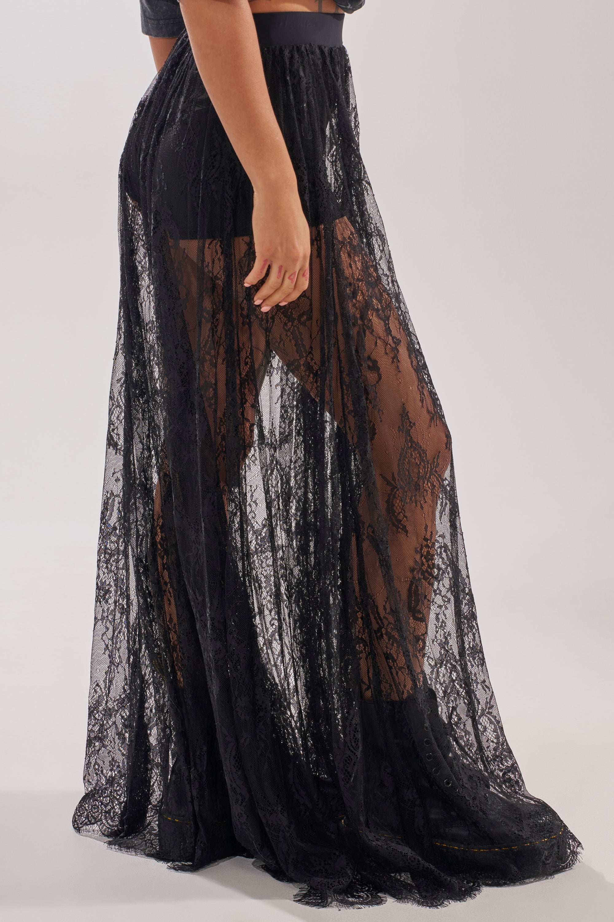 A person wears the LOVELY DAYS LACE MAXI SKIRT—a sheer black lace maxi over black shorts and boots. The floor-length lace reveals their legs as they stand sideways, making it a chic choice for resort wear. 1