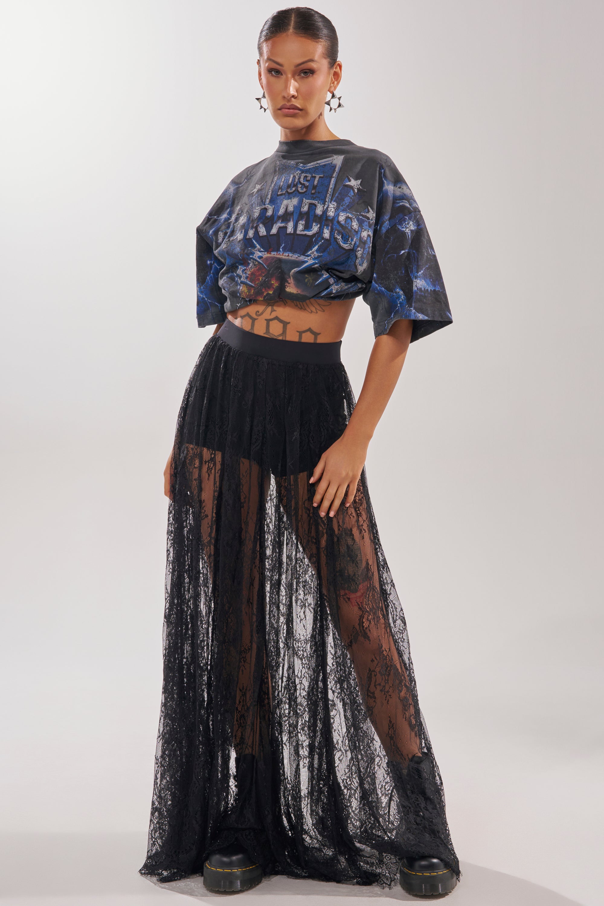 A woman stands confidently in chic resort wear, featuring the LOVELY DAYS LACE MAXI SKIRT over high-waisted shorts, a cropped graphic t-shirt, black boots, and hoop earrings against a plain light background. 3