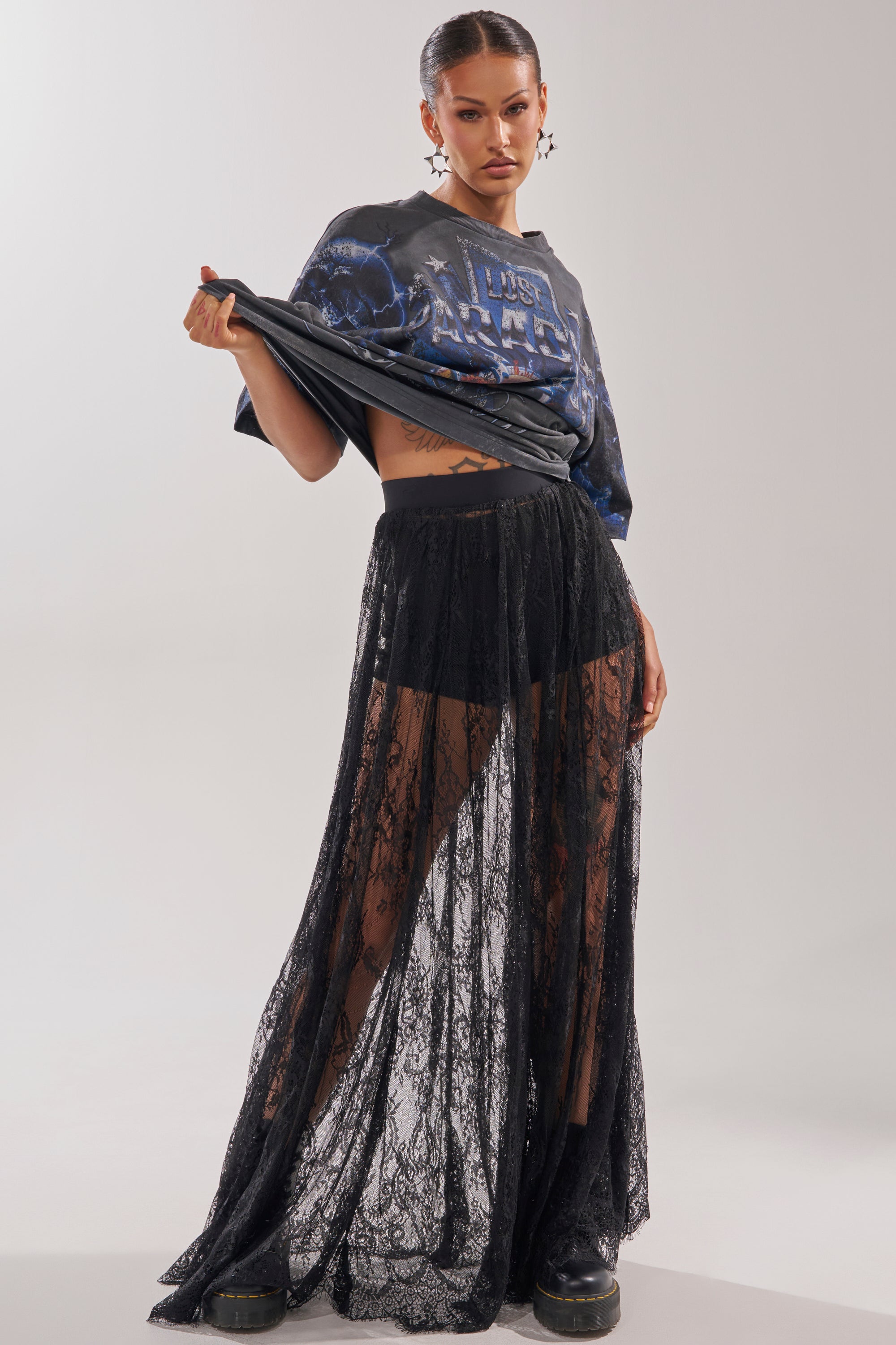 A woman poses confidently, lifting her graphic tee to reveal her midriff. She pairs sleek hair and statement earrings with the LOVELY DAYS LACE MAXI SKIRT over black shorts and chunky black boots for resort-ready flair. 5
