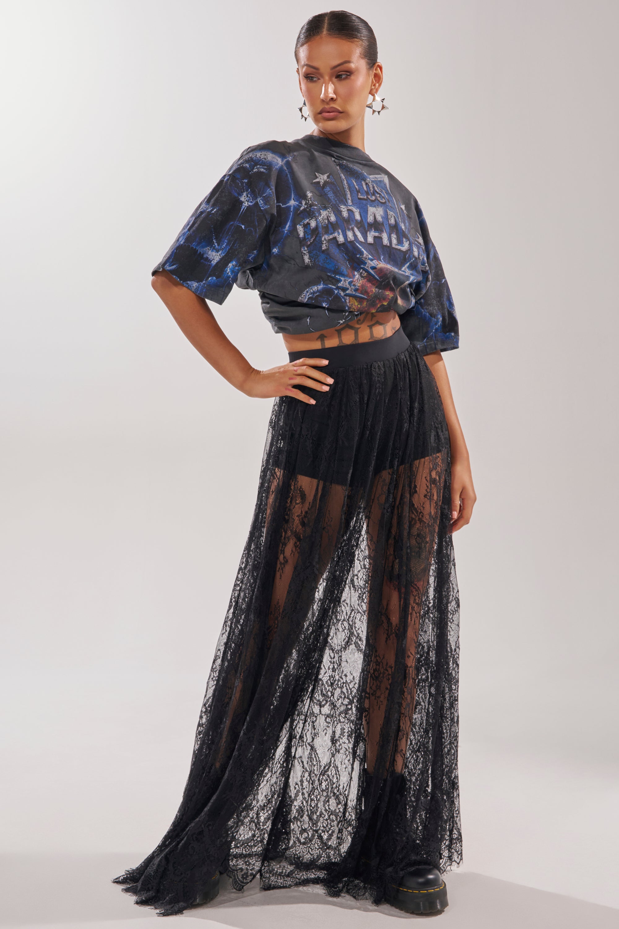 A woman models the LOVELY DAYS LACE MAXI SKIRT layered over black shorts, styled with a graphic cropped tee and chunky black shoes. Her slicked-back hair and star earrings add an edgy touch to this resort-inspired look. 6
