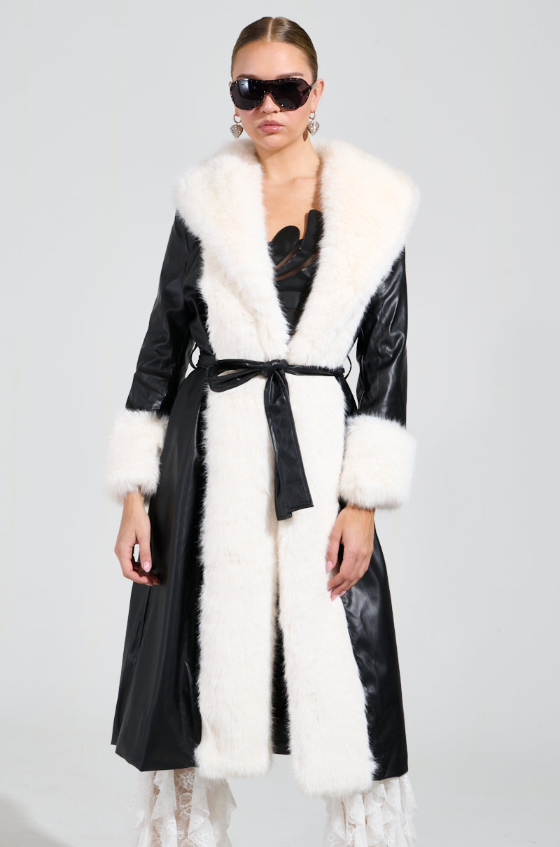 A person dressed in the stylish CANT GET ENOUGH FUR TRENCH, adorned with white trim and oversized sunglasses, poses against a simple background. The winter coat is highlighted with lace accents and elegantly tied at the waist with a belt.