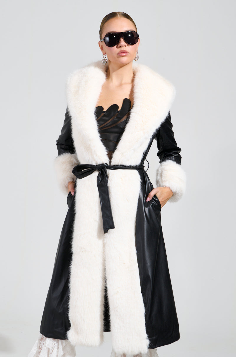 A person confidently poses in the CANT GET ENOUGH FUR TRENCH, a long black faux fur coat with white trim, accompanied by black gloves and sunglasses. The coat is belted at the waist, with hands tucked casually in its pockets against a plain gray backdrop.