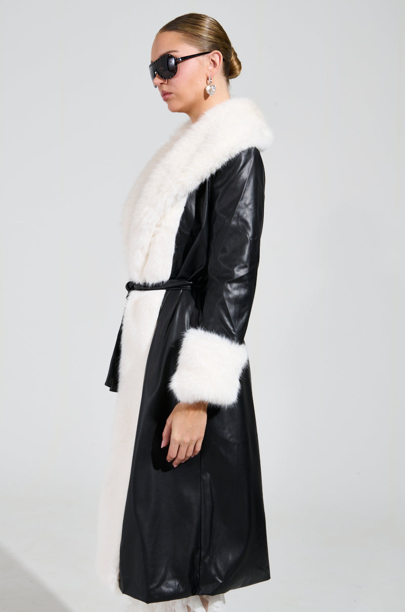 A person wearing sunglasses models the chic "CANT GET ENOUGH FUR TRENCH," a black faux leather coat featuring white faux fur trim and cuffs. This stylish winter jacket is belted at the waist, adding elegance to the look. The plain background perfectly highlights this fashionable ensemble.