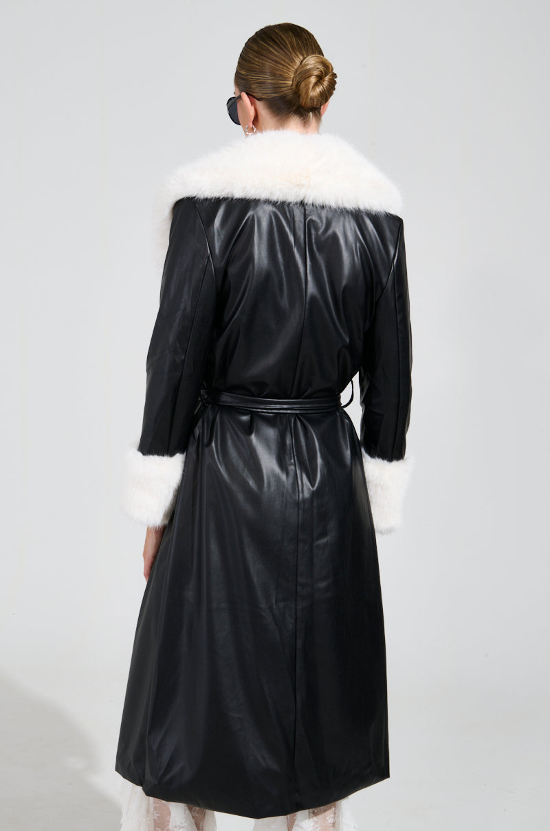 Wearing the CANT GET ENOUGH FUR TRENCH, an individual is pictured from behind in a stylish long black faux leather coat featuring white faux fur trim on the collar and cuffs. With their hair styled up and sunglasses accentuating their look, the plain background underscores this chic winter jacket ensemble.