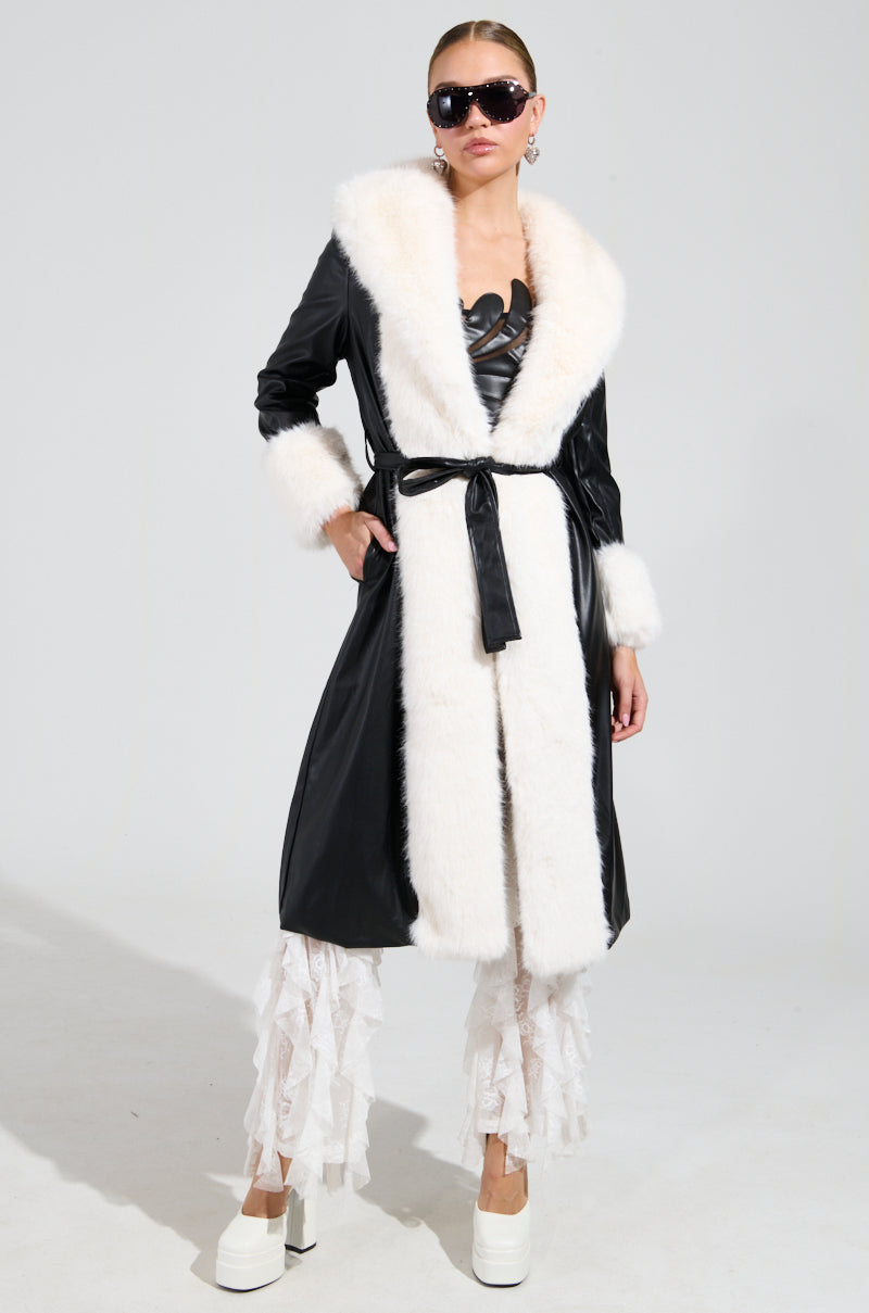 Wearing the striking "Can't Get Enough Fur Trench," a person poses confidently in sunglasses. They pair this statement faux leather trench, featuring white fur trim, with chic white ruffled pants and stylish white platform shoes against a plain background.