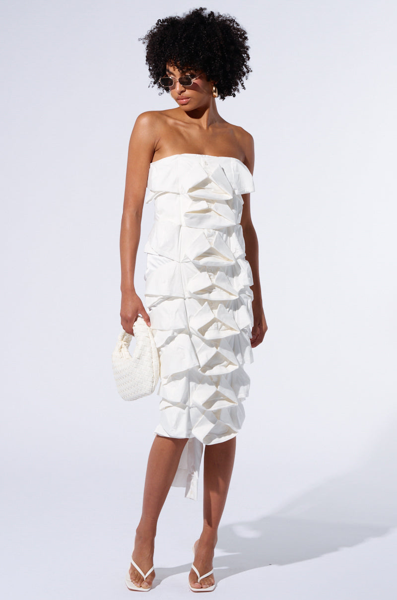 A woman with curly black hair dons the POPPIN MY COLLAR LAYERED MIDI DRESS, featuring a strapless neckline and a textured white fabric. She complements her look with round sunglasses, white sandals, and a stylish white woven handbag. The backdrop is plain white.