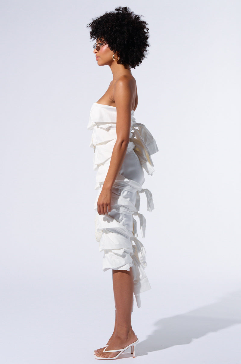 A woman with curly hair stands in profile, wearing the "Poppin My Collar Layered Midi Dress," which features a strapless neckline and is adorned with multiple large bows along its length. She is also wearing white heeled sandals, set against a plain white background.