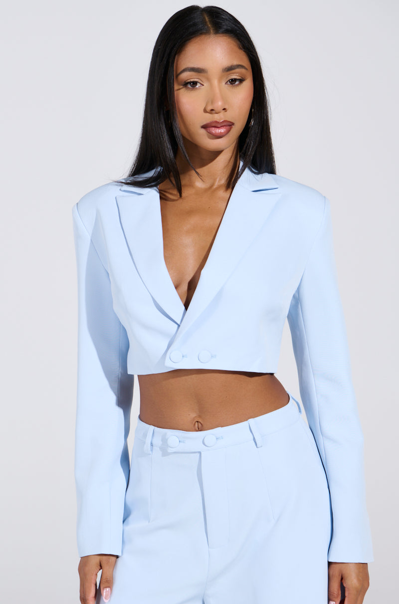 A woman with long black hair wears the LOTTIE CROPPED BLAZER IN LIGHT BLUE from the AKIRA Label, paired with matching high-waisted trousers. The blazer features a deep V-neck and button detail, complementing her confident expression against a plain white background.