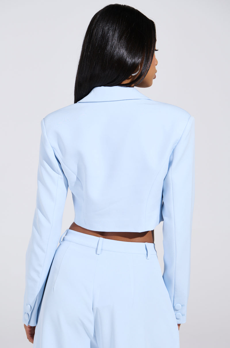 Against a plain background, someone with long dark hair is shown from the back wearing a chic LOTTIE Cropped Blazer in light blue with matching high-waisted trousers.