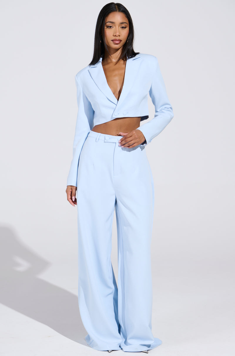 A person confidently wears the AKIRA Label's LOTTIE CROPPED BLAZER in light blue, paired with high-waisted, wide-leg pants. Their long dark hair cascades over their shoulders against a plain backdrop.