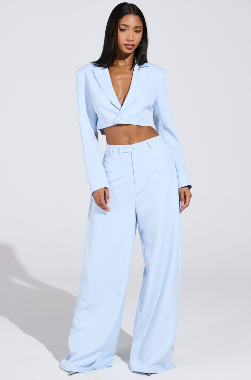 In a LOTTIE CROPPED BLAZER IN LIGHT BLUE set, a woman exudes confidence with high-waisted, wide-leg trousers. Her sleek straight black hair and minimal accessories enhance the ensemble's elegance against a simple backdrop.