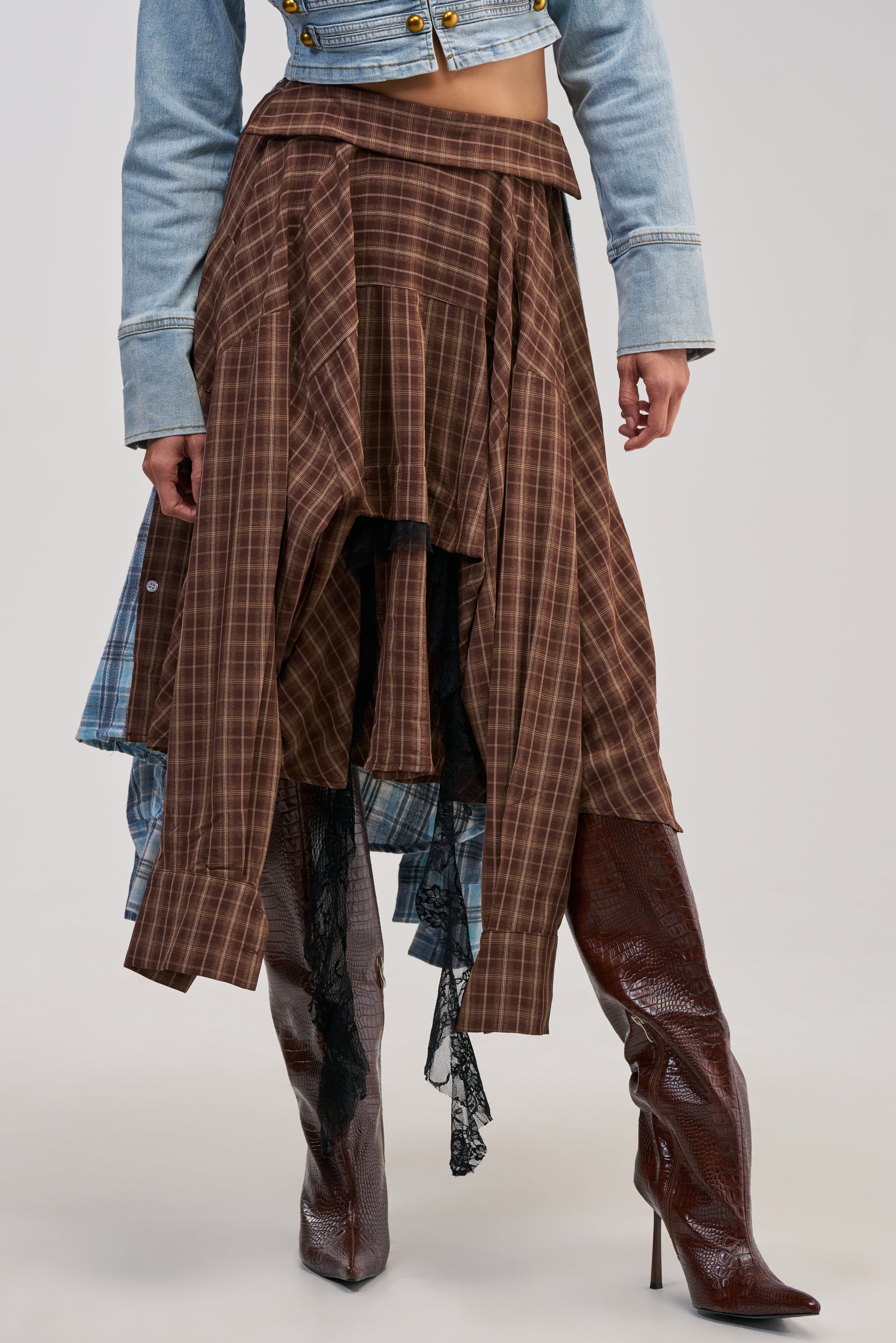 A person wears a cropped denim jacket, the FEELING ALL THE VIBES PLAID SKIRT with its asymmetrical midi silhouette and layered fabric for statement styling, and tall, brown textured boots.