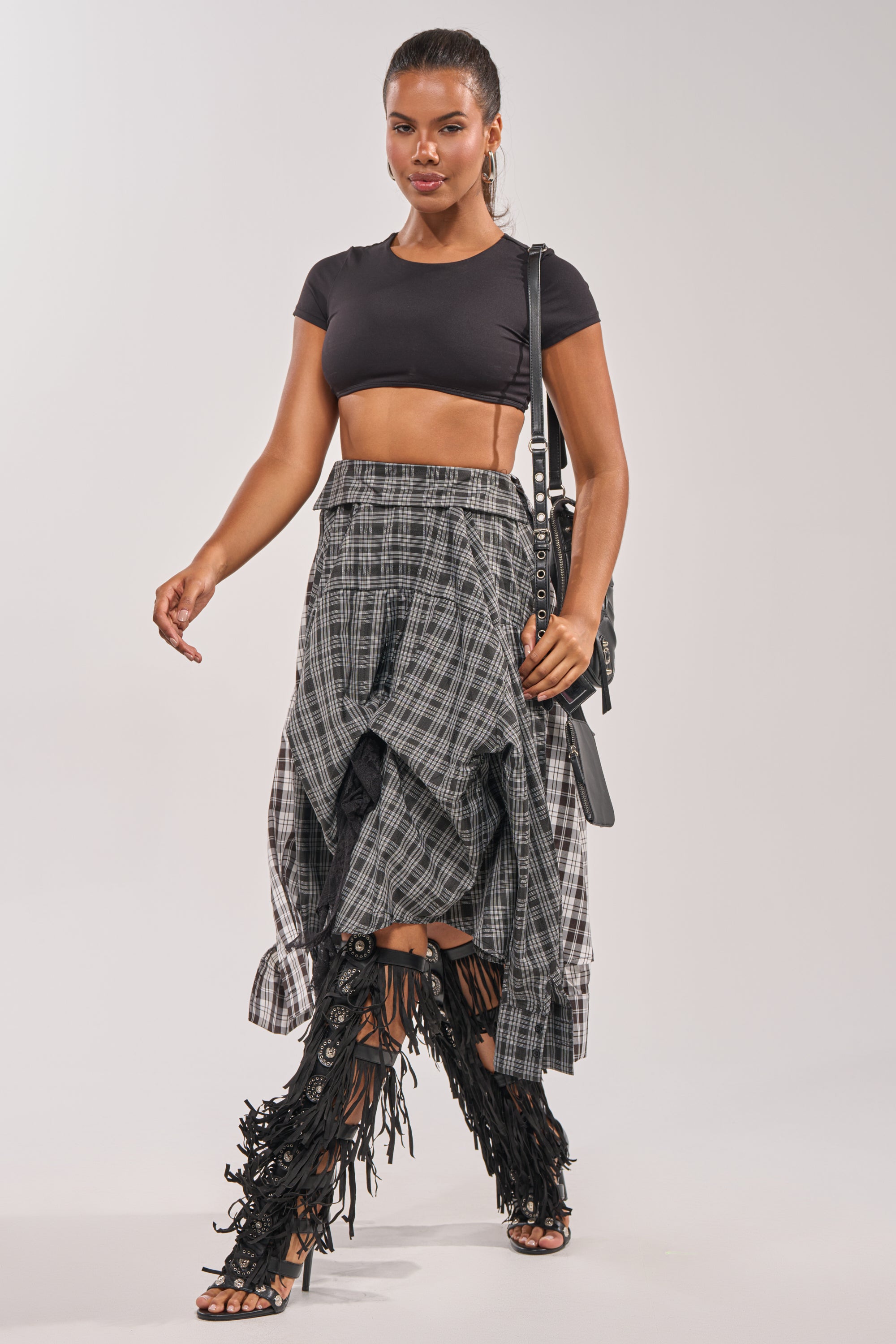 A woman walks confidently against a plain light background, wearing the FALL VIBES PLAID MIDI SKIRT, a black crop top, black fringe knee-high heels, and carrying a black bag. 2