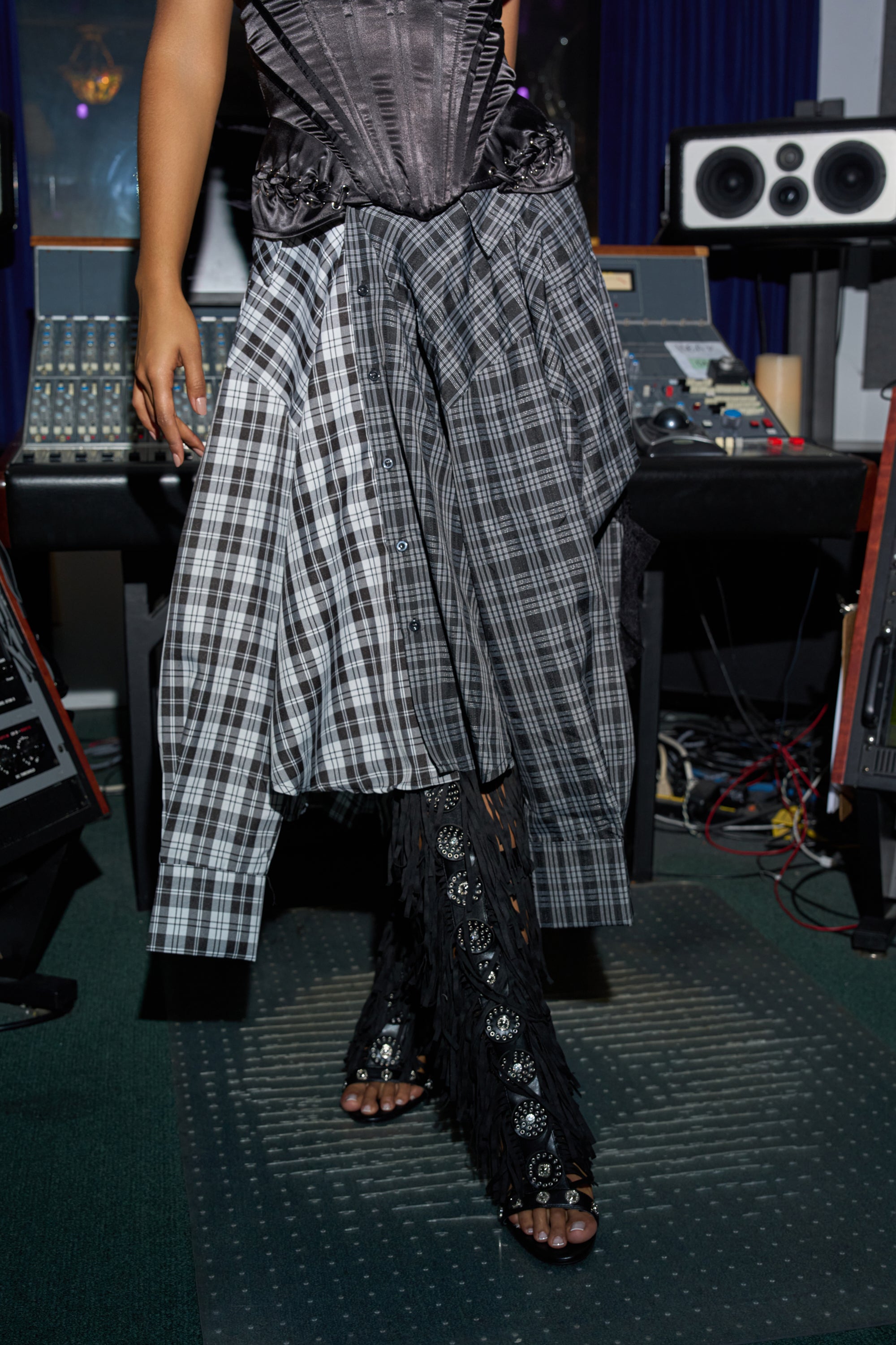 A person wearing the FALL VIBES PLAID MIDI SKIRT, a black corset, fringe leg warmers, and jeweled sandals stands near audio equipment. 1