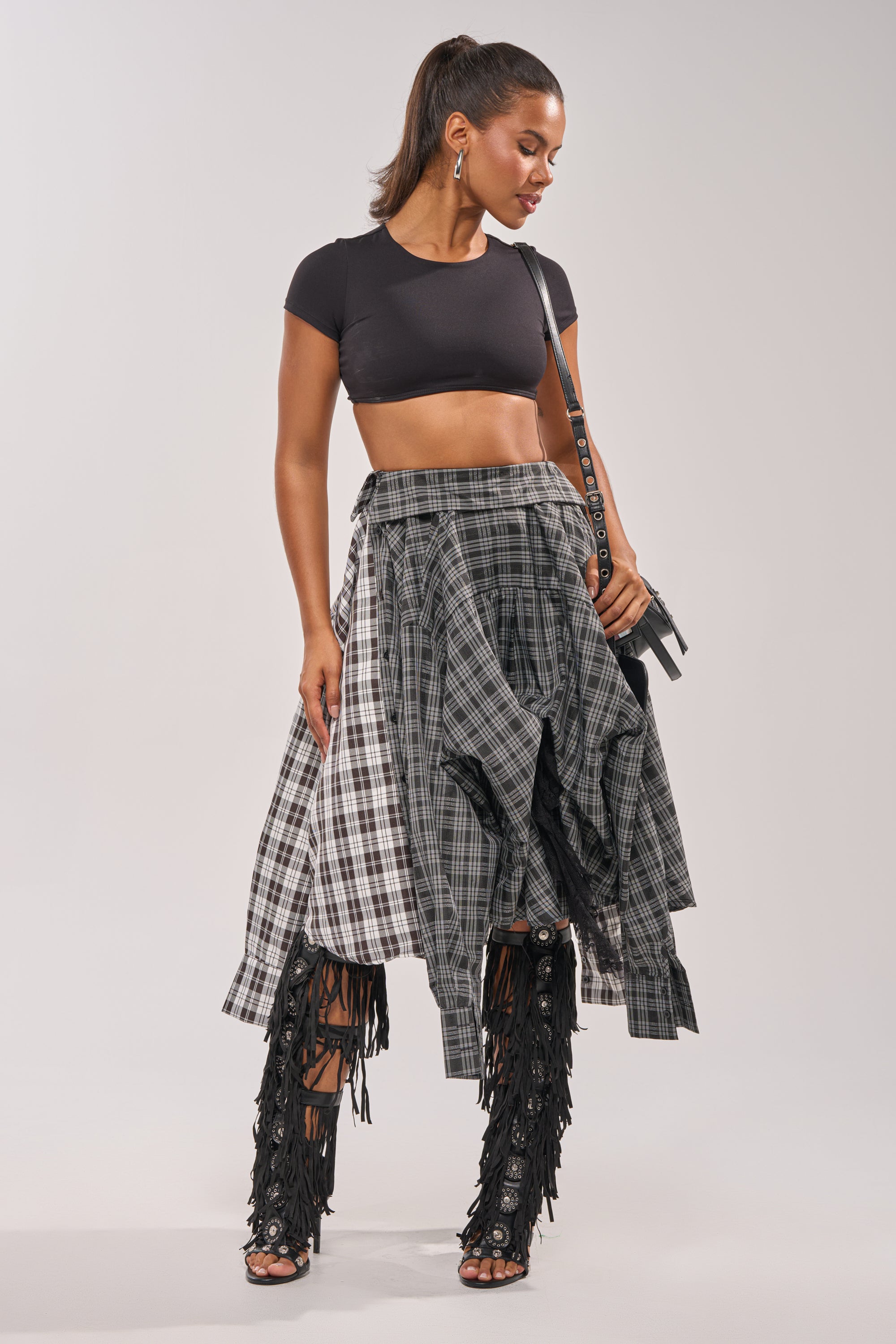 A woman in a black crop top, FALL VIBES PLAID MIDI SKIRT, black fringe heeled boots, and a small black bag stands against a plain light background, looking down with a slight smile. 8