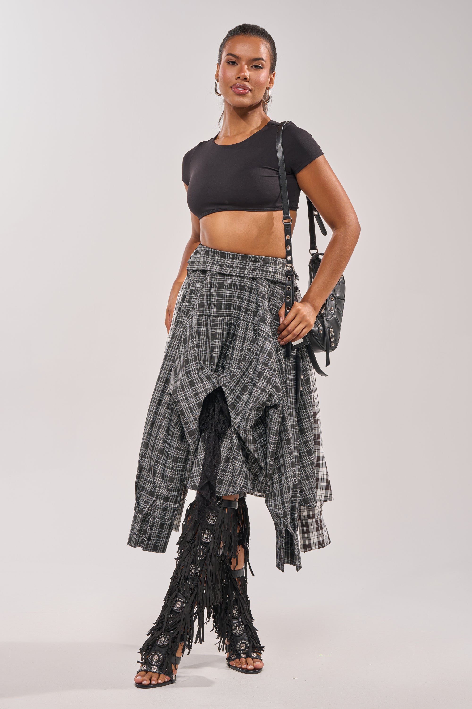 A woman confidently poses against a plain background wearing the FALL VIBES PLAID MIDI SKIRT, a black crop top, knee-high fringe boots, and a black shoulder bag. 9