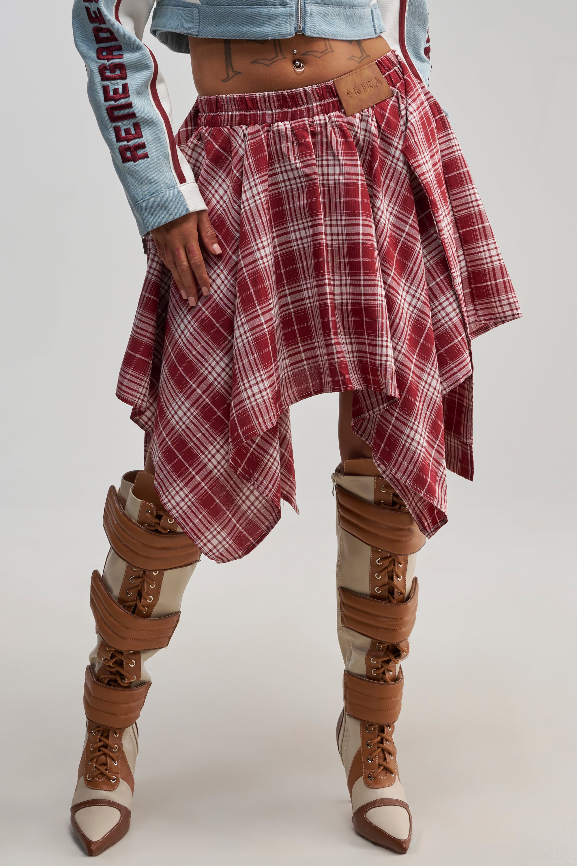Person wearing the TIME FOR SOME FUN PLAID MINI SKIRT in red, a denim jacket with “RENEGADE” on the sleeve, and tan platform boots with brown straps against a plain background.