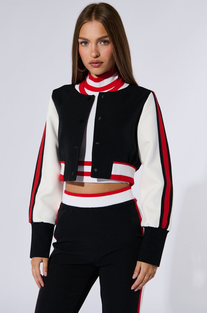 A woman with long brown hair stands against a white background, wearing an AKIRA Label RAYE CROPPED BOMBER featuring a color block pattern with red accents. She also wears matching black and white pants with red stripes down the sides and a high-neck striped top.