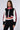 A woman with long brown hair stands against a white background, wearing an AKIRA Label RAYE CROPPED BOMBER featuring a color block pattern with red accents. She also wears matching black and white pants with red stripes down the sides and a high-neck striped top.