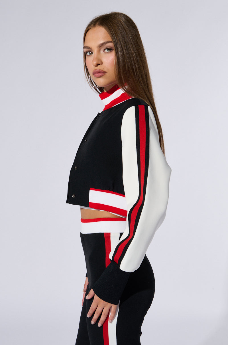 A woman with long brown hair stands against a plain background wearing a sporty black, white, and red AKIRA Label RAYE CROPPED BOMBER with matching pants. The jacket features red and white stripes along the sleeves, and the pants have coordinating side stripes. She is looking at the camera.