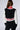 A person with long brown hair is seen from behind, wearing the RAYE CROPPED BOMBER from AKIRA Label. The sleek knit fabrication showcases a black body, white sleeves, and red-and-white striped cuffs and waistband in a fashionable color-block pattern. They are also wearing black pants.