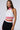 A person with long brown hair is wearing an AKIRA Label RAYE CROPPED HALTER TOP in white with red stripes, paired with black high-waisted pants featuring a contrast striped pattern. This sporty chic ensemble stands out against the plain white background.