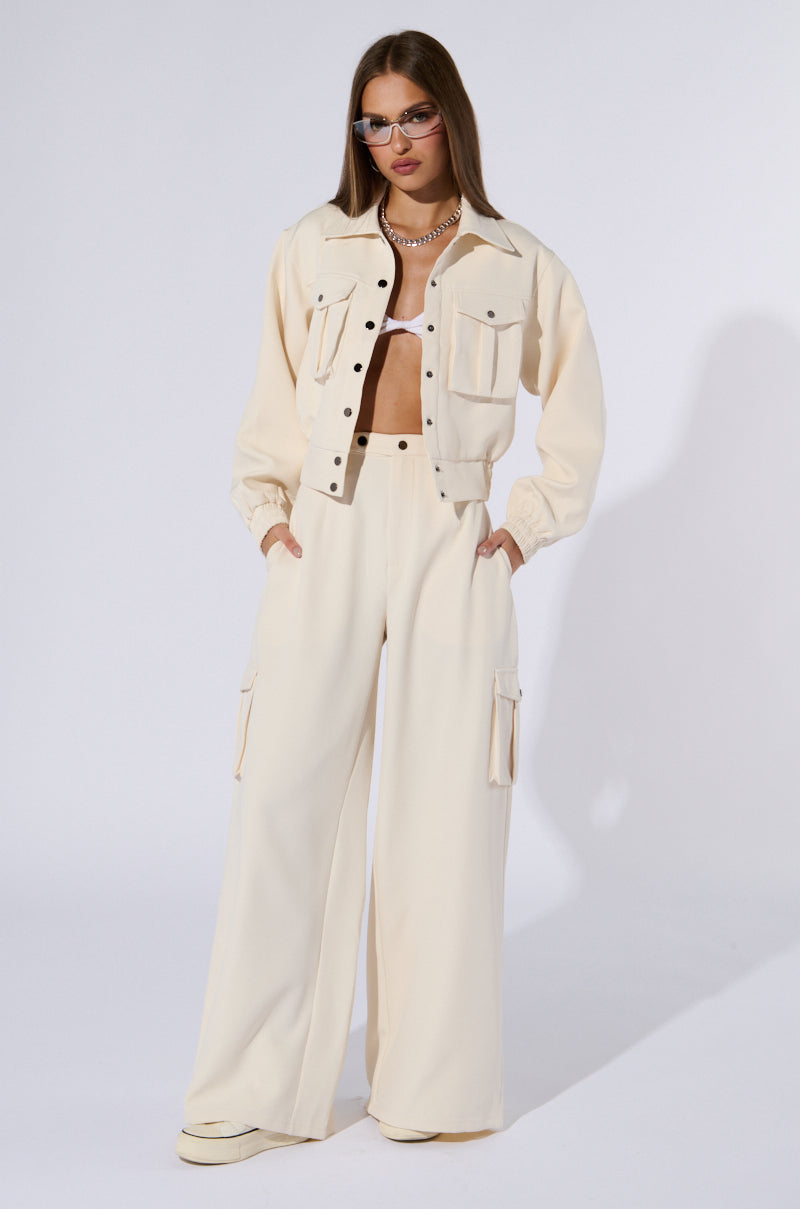 A woman stands dressed in a chic cream-colored ensemble, featuring a button-up jacket, a cropped white top, and the stylish BUTTERCUP CARGO TROUSER. She accessorizes with sunglasses and white shoes, has long loose hair cascading over her shoulders, and her hands rest casually in her pockets. The plain white background behind her casts a faint shadow.