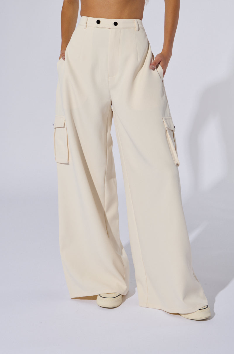 A person dressed in the BUTTERCUP CARGO TROUSER, which are high-rise wide-leg cream trousers with cargo-inspired pockets on each side, stands with their hands in their pockets. The trousers showcase black button details at the waist, and white shoes are partially visible at the bottom.