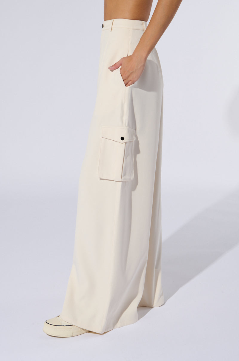 A person wearing the BUTTERCUP CARGO TROUSER with a cargo-inspired pocket poses with one hand in the pocket. The high-rise, wide-leg trousers drape loosely to the ground, covering most of the white sneakers, against a plain white background.
