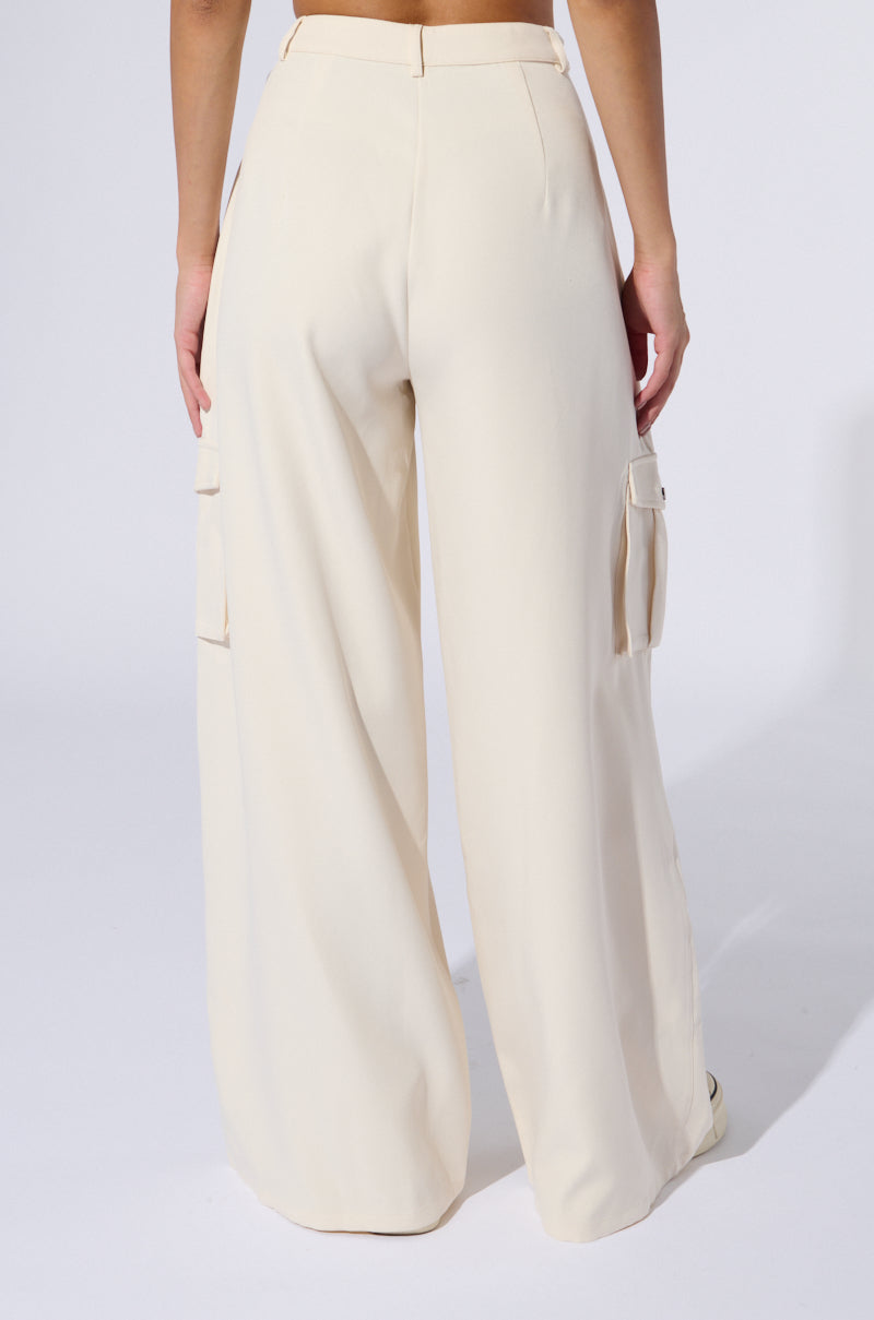 A person is wearing the BUTTERCUP CARGO TROUSER, which features high-rise wide-leg trousers in a beige hue and cargo-inspired pockets on the sides. The image, taken from the back, showcases the full length of the pants and a portion of the person's midriff and hand against a plain, light-colored background.