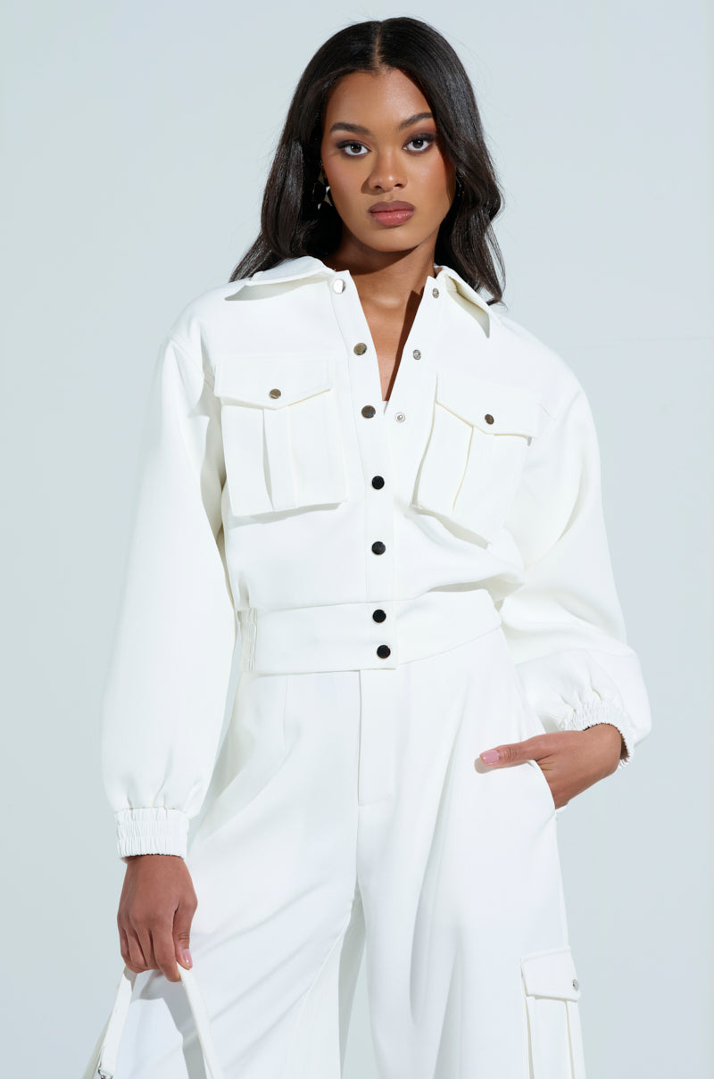 A woman stands confidently in the Buttercup Cargo Bomber in white, featuring black buttons, large chest pockets, and matching wide-leg pants. Her long dark hair drapes over the jacket as she maintains a neutral expression against a light background.