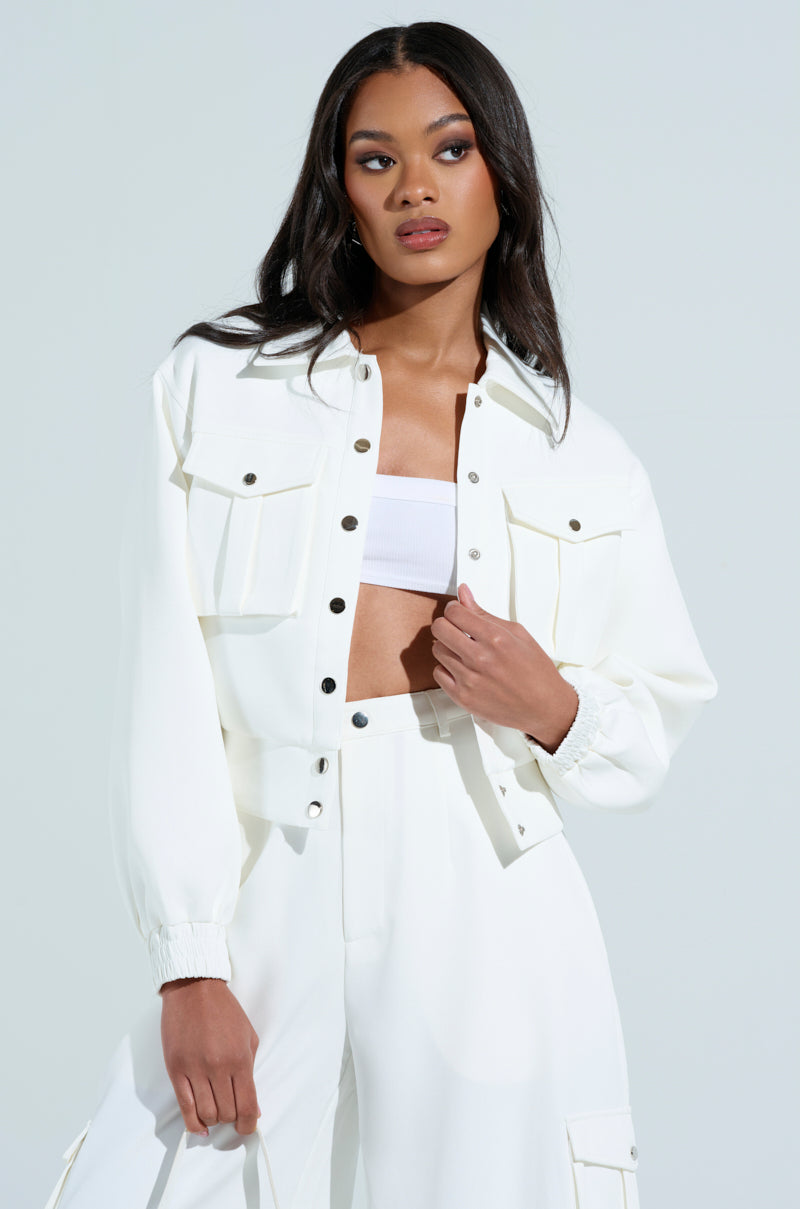 A woman in a BUTTERCUP CARGO BOMBER IN WHITE and high-waisted pants stands against a plain background. The jacket features front pockets and snap buttons. Her long dark hair frames her face as she gazes to the side with a neutral expression.