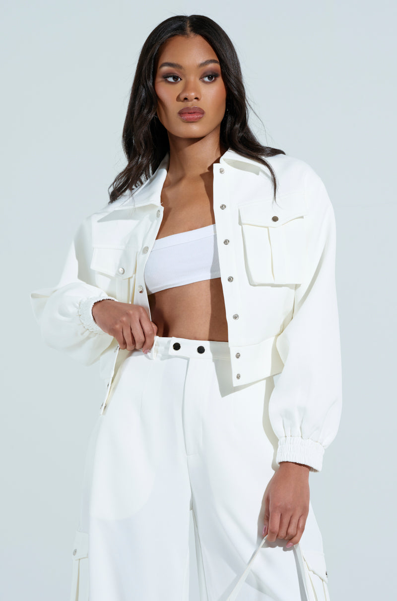 The woman wears the chic AKIRA Label BUTTERCUP CARGO BOMBER IN WHITE with pockets, paired with high-waisted pants and a white crop top, set against a light background.