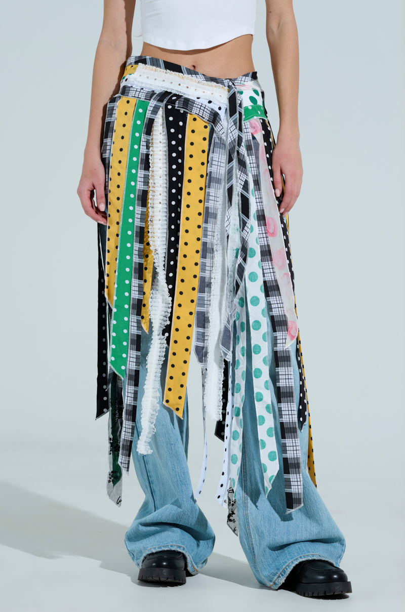 A person wears a white crop top, patchwork jeans, and the OH SO CHIC PATTERN MIX BELT SKIRT adorned with stripes, polka dots, and plaid patterns. The fringed skirt has a dangling tassel design. They're also in black platform shoes.