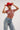A person confidently wears a red cowboy hat, the BEJEWELED RUBY SLEEVELESS TOP with large red and silver links, and blue jeans featuring metallic chain links, against a plain white backdrop.