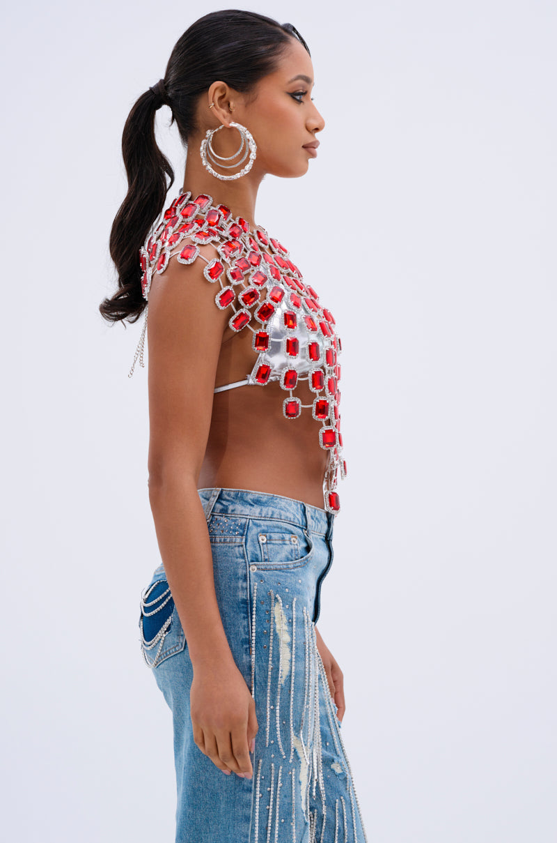 A woman with long hair tied back is wearing a BEJEWELED RUBY SLEEVELESS TOP adorned with red and faux silver geometric shapes, paired with painted blue jeans. In profile, she flaunts large hoop earrings against a plain background.