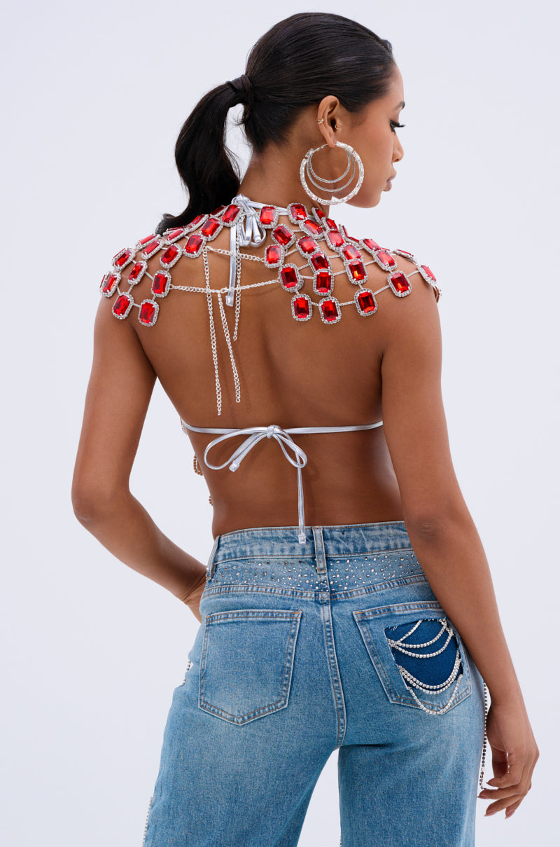 A woman with dark hair tied back models the BEJEWELED RUBY SLEEVELESS TOP, featuring red gem decorations and metallic chain links. She pairs it with large hoop earrings and chain-embellished blue jeans, posing elegantly against a plain white background.