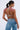 A woman with dark hair tied back models the BEJEWELED RUBY SLEEVELESS TOP, featuring red gem decorations and metallic chain links. She pairs it with large hoop earrings and chain-embellished blue jeans, posing elegantly against a plain white background.