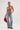 A woman in a red cowboy hat stands confidently, her BEJEWELED RUBY SLEEVELESS TOP shimmering with rhinestones. She holds the hat by her side, wearing distressed jeans with fringe and metallic chains. Large earrings and matching red boots complete her bold look.