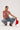 A person kneels wearing a BEJEWELED RUBY SLEEVELESS TOP with silver and red beads, denim fringe pants, red cowboy boots, and a metallic chain accessory. They hold a red hat on a white background, highlighting the vibrant details.
