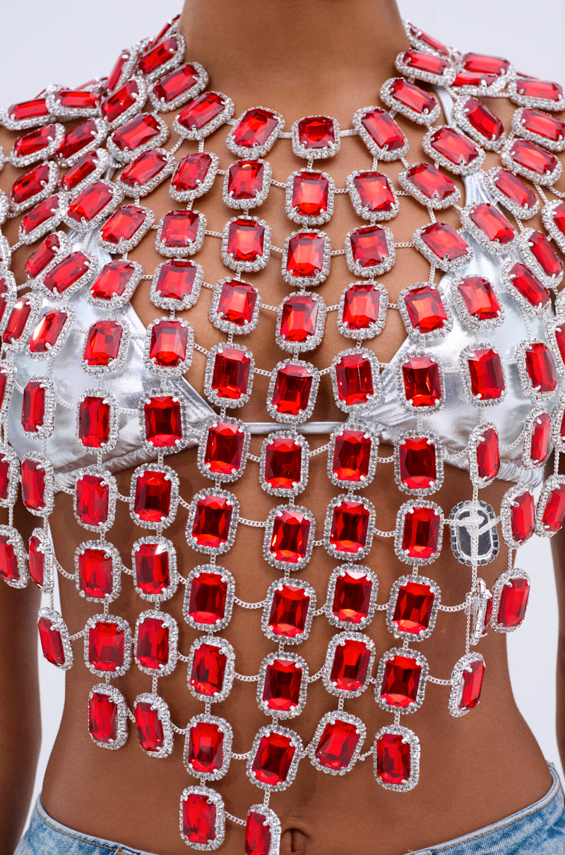 A close-up shows a person wearing the BEJEWELED RUBY SLEEVELESS TOP, adorned with red, rectangular jewels connected by metallic chains over a metallic silver bra, paired stylishly with blue jeans against a simple white backdrop.