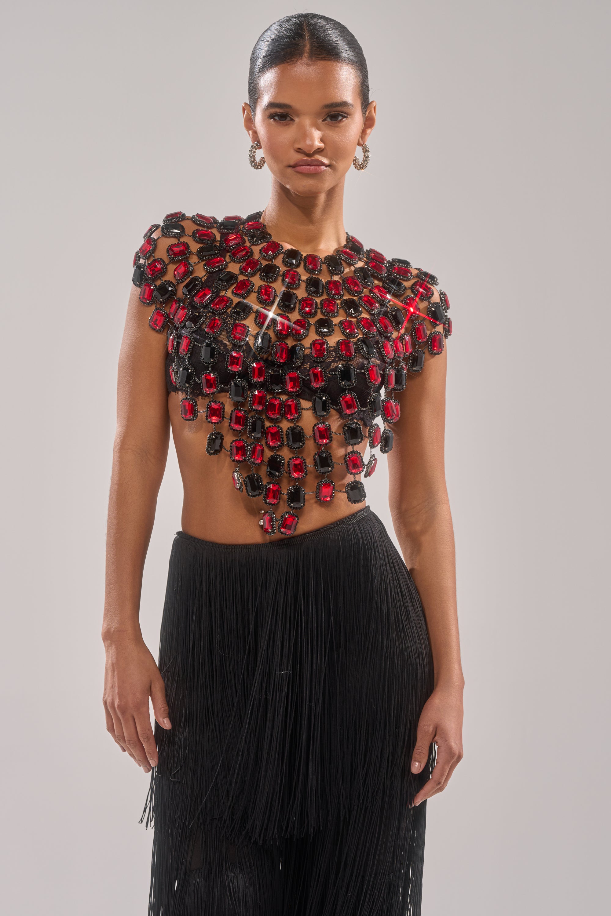 A woman poses confidently in the RUBY RHINESTONE BLOUSE, adorned with large red and black stones. The midriff-baring top pairs with a black fringed skirt, set against a plain background that highlights the blouse's bold gemstone design. 0