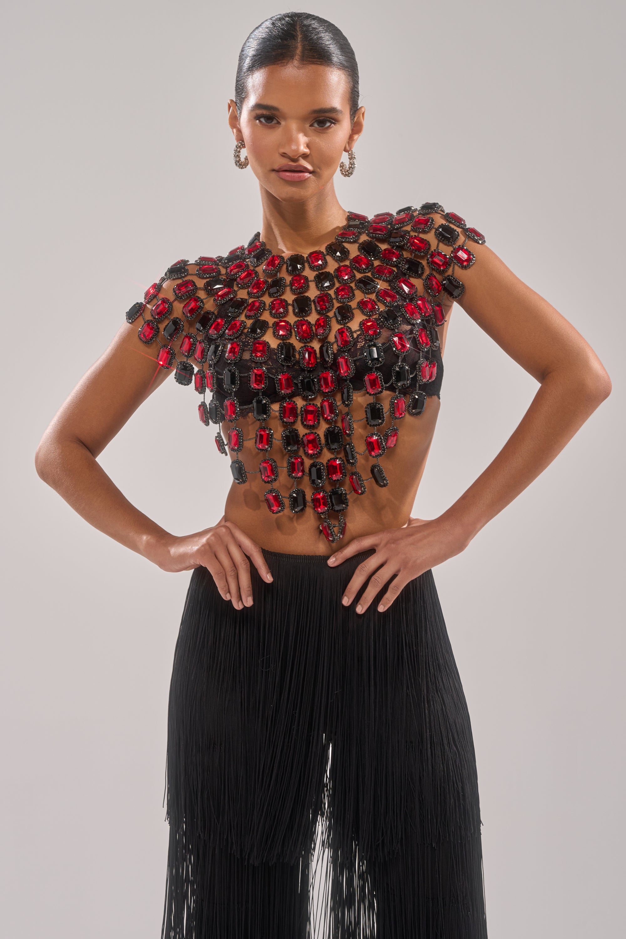 A person wears the RUBY RHINESTONE BLOUSE, a bold chain link top with large red and black stones, paired with black fringed pants, standing hands on hips against a plain background. 1