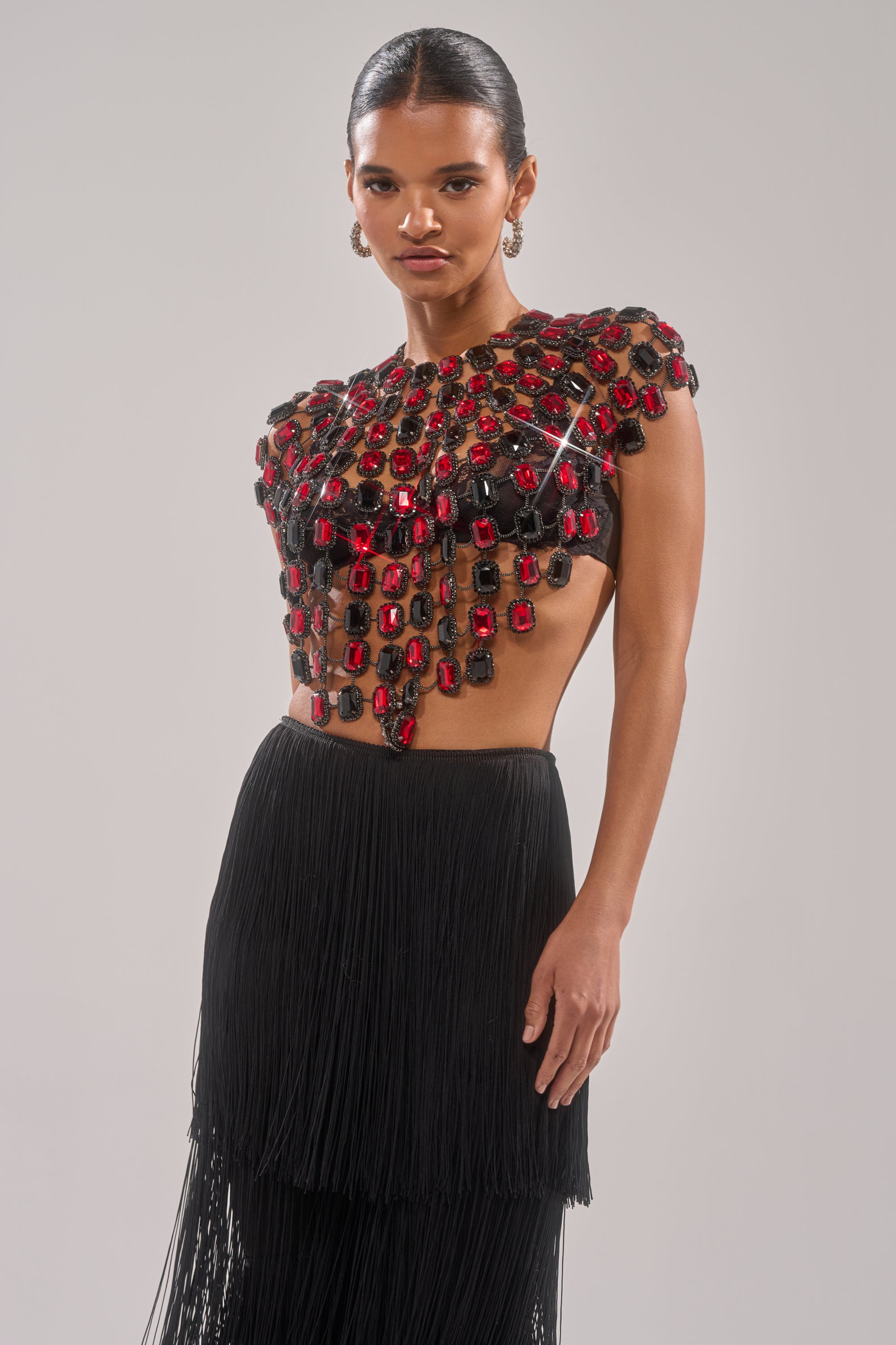 A woman poses in the RUBY RHINESTONE BLOUSE, featuring bold red and black stones, an open midriff, and a high-waisted black fringed skirt against a plain background. 2