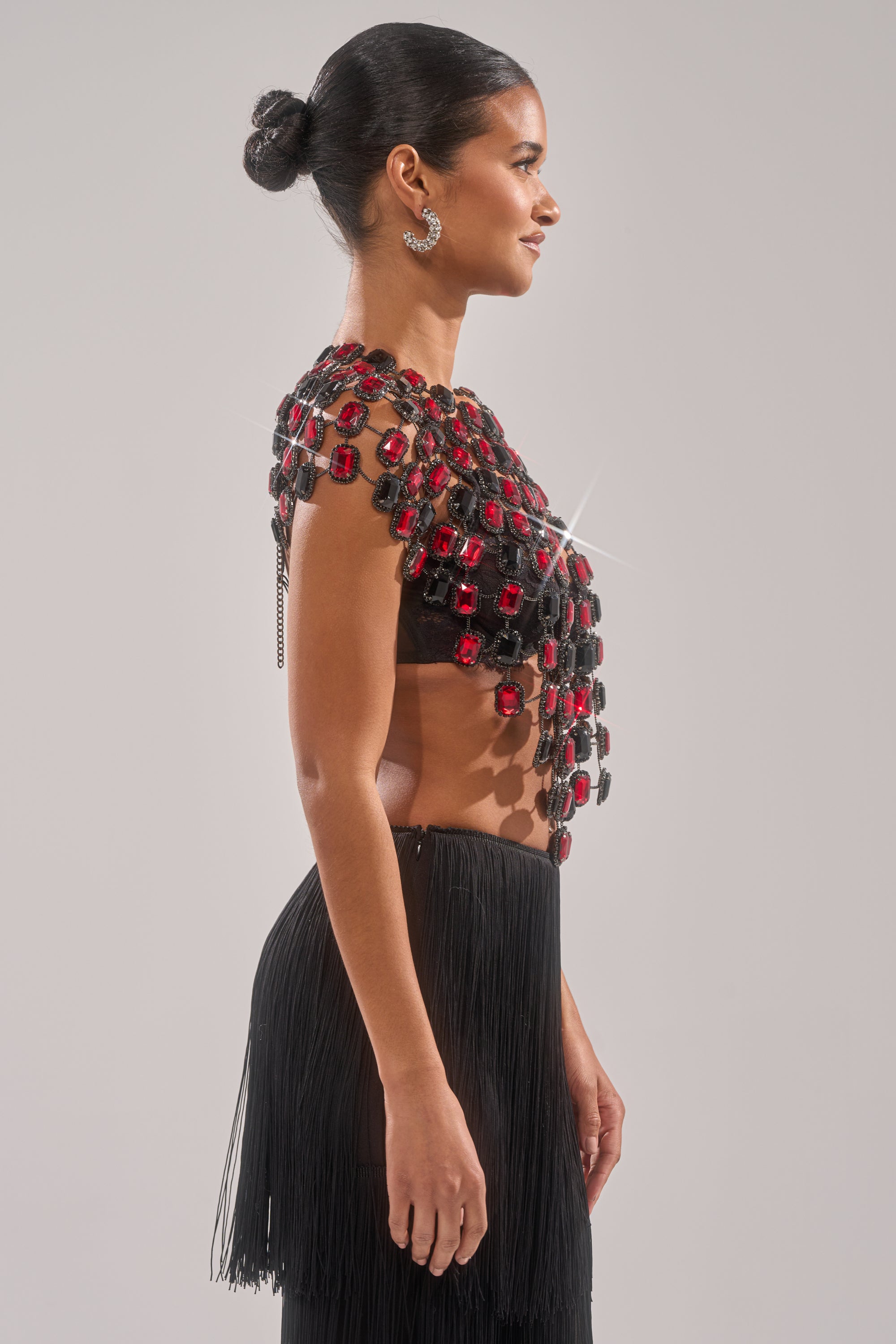 A woman in profile wears the RUBY RHINESTONE BLOUSE, a black crop top with gemstone detailing, paired with a long black fringe skirt. Her hair is in a sleek bun, and she accessorizes with silver earrings. 3