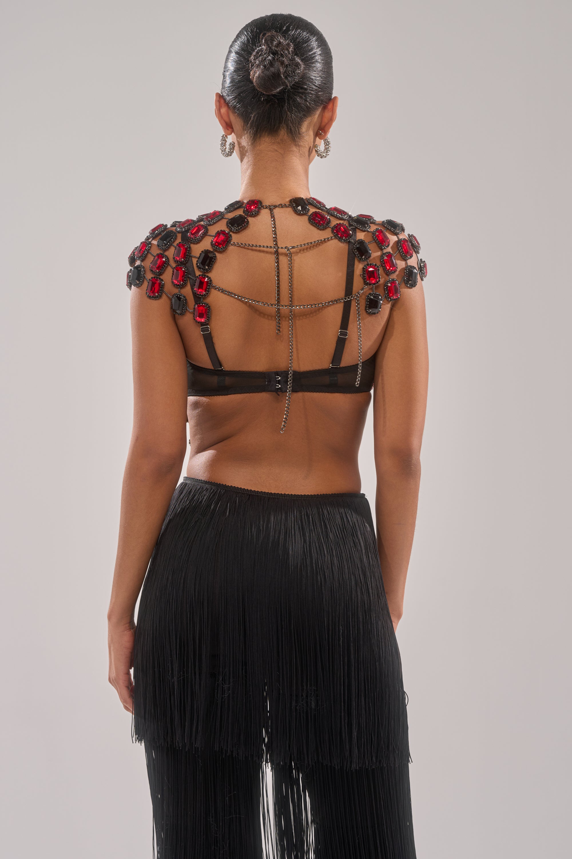 A woman stands facing away, wearing a black skirt with long fringes and the RUBY RHINESTONE BLOUSE, a chain link top with gemstone details. Her hair is in a neat bun, set against a plain light gray background. 4