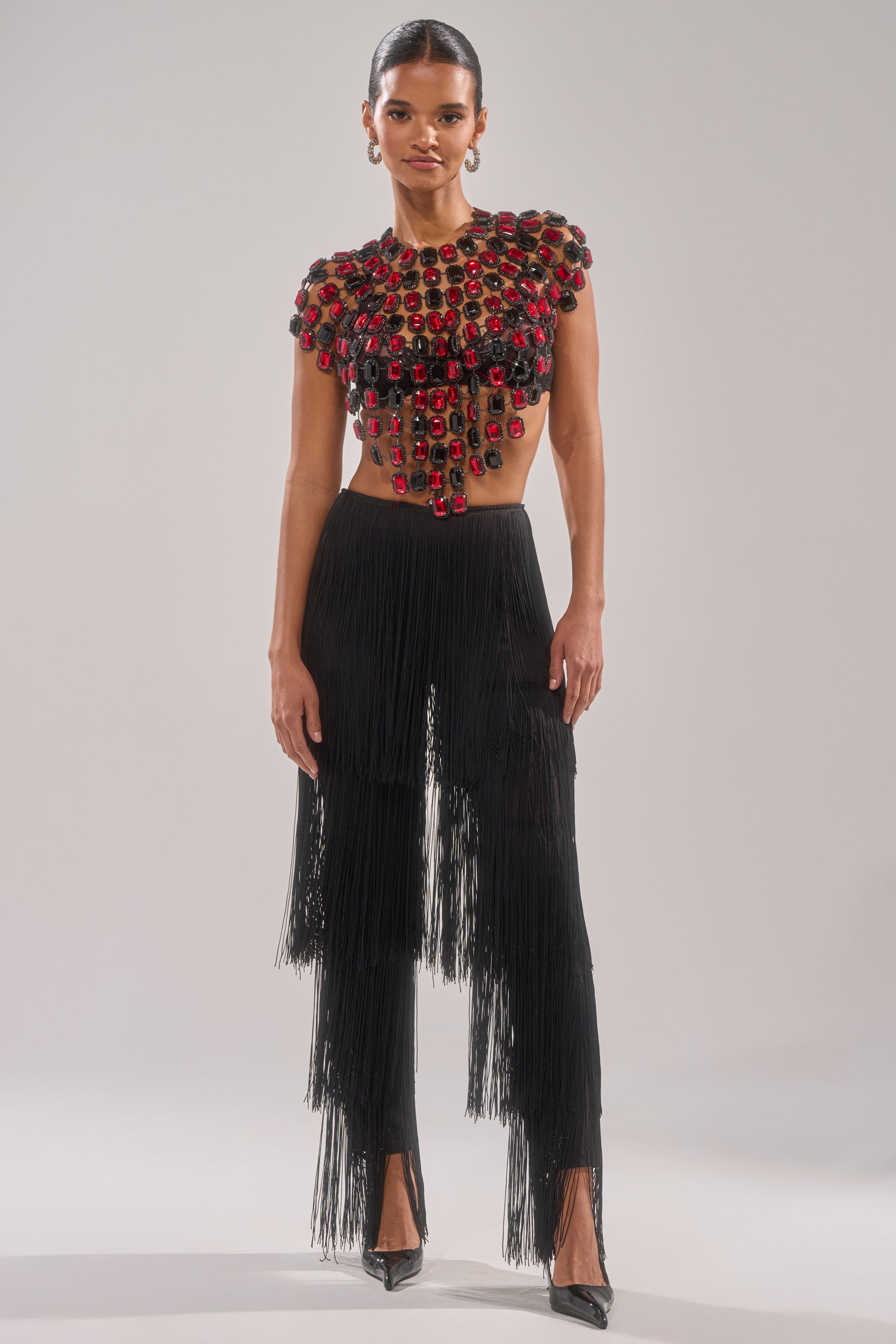 A woman exudes confidence in the RUBY RHINESTONE BLOUSE paired with black fringe pants. Red and black beading sparkles on the cropped top, while her slicked-back hair and black heels finish the chic look against a plain background. 5